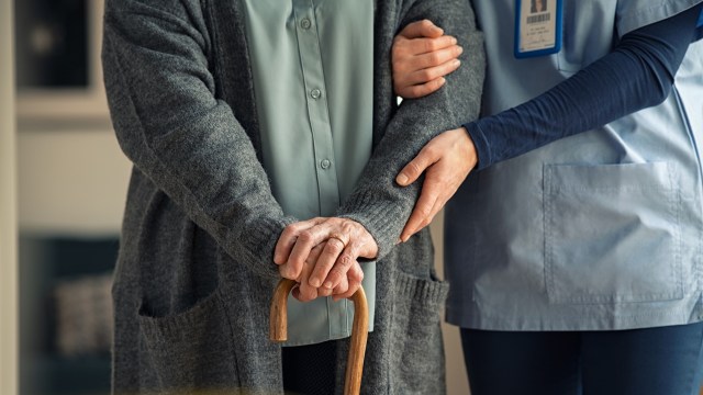 An older person holds a cane while a caregiver gently supports their arm.