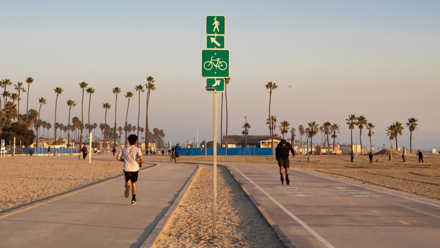 A pedestrian and a person on rollerblades along a beachside walkway with palm trees and signs for walking and biking.