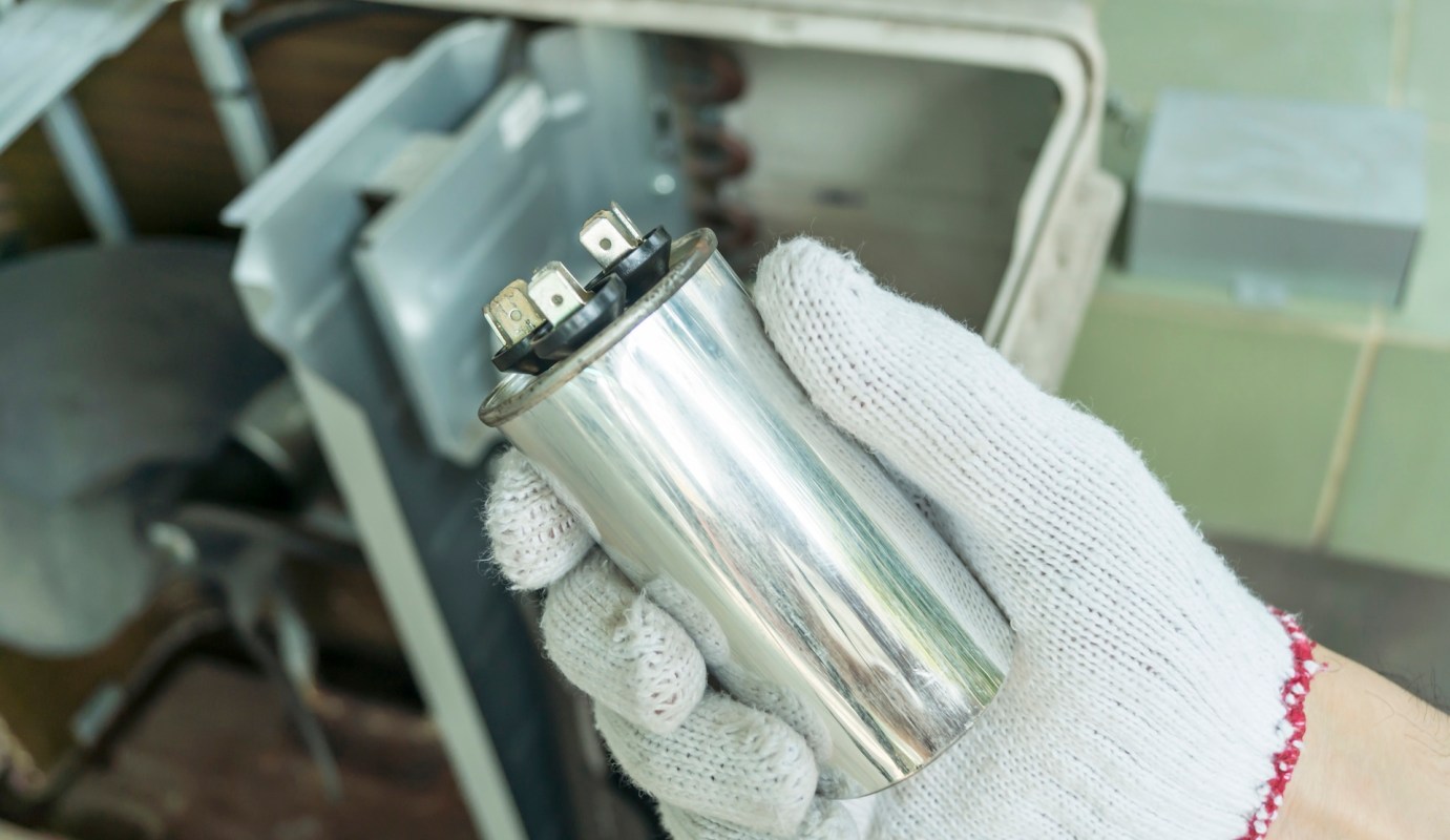 A person wearing a glove holds a silver capacitor with electrical terminals.