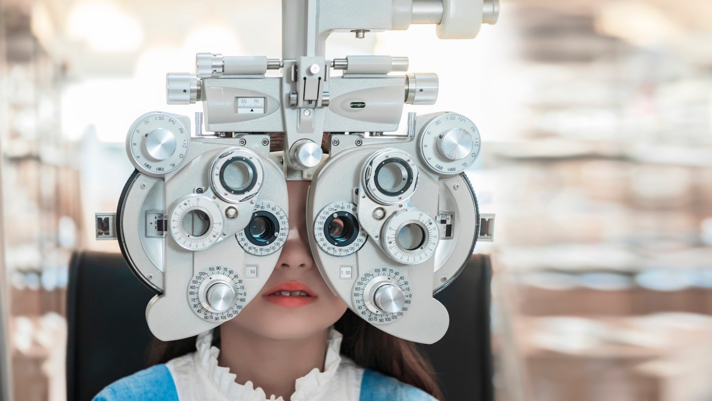 A child behind an eye test machine in a clinical setting with a blurred background.