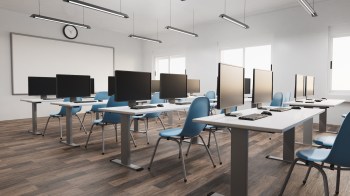An empty classroom with several computer desks, blue chairs, and large windows providing natural light.