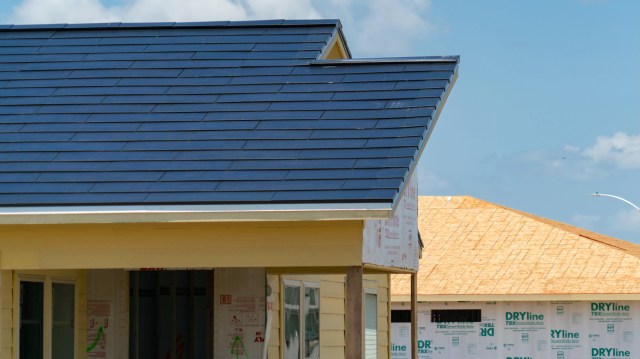 A house under construction with Tesla Solar Roof panels and an adjacent unfinished structure.