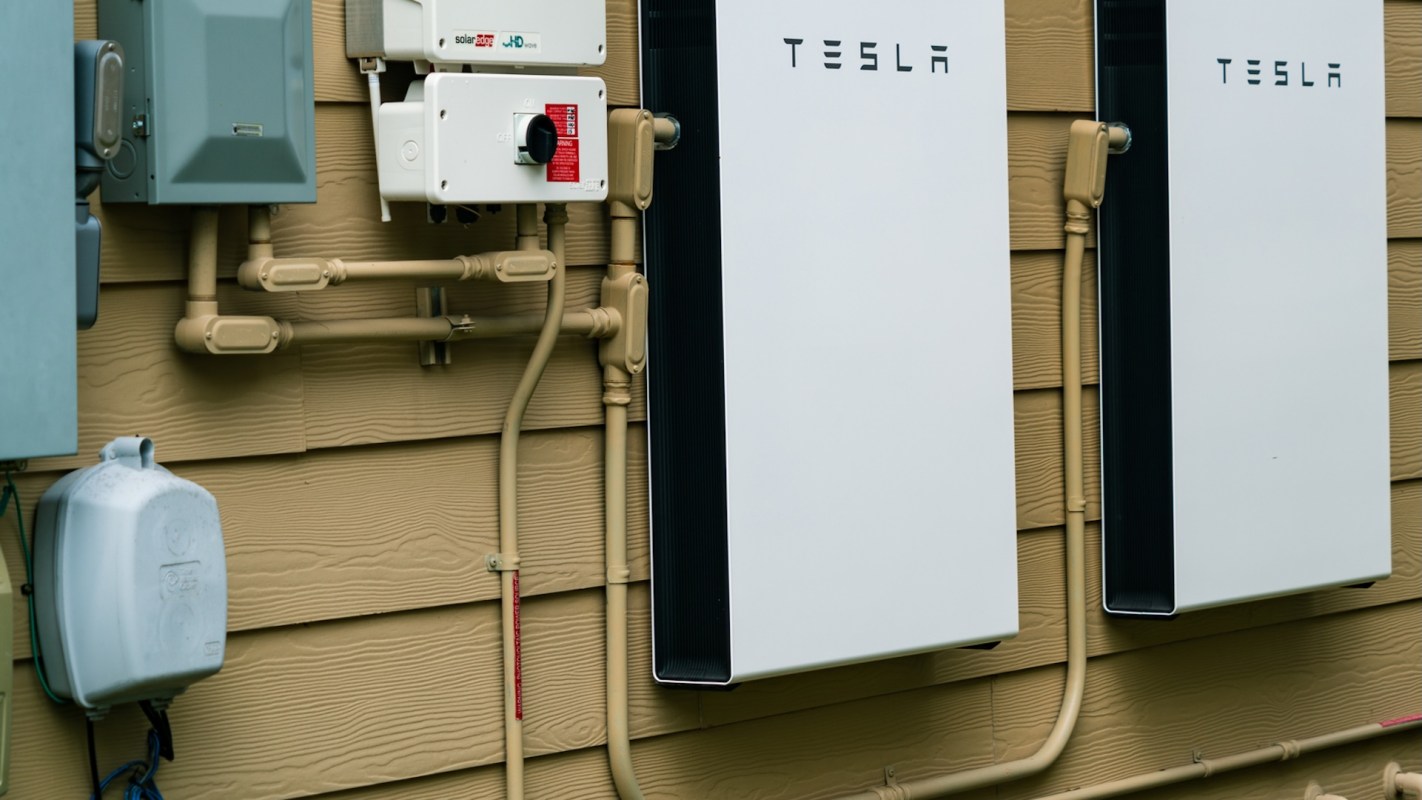 Two Tesla Powerwall units mounted on a house exterior near electrical equipment and piping.