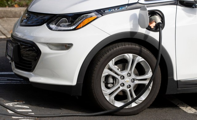 A white Chevrolet Bolt electric vehicle is plugged into a charging station.