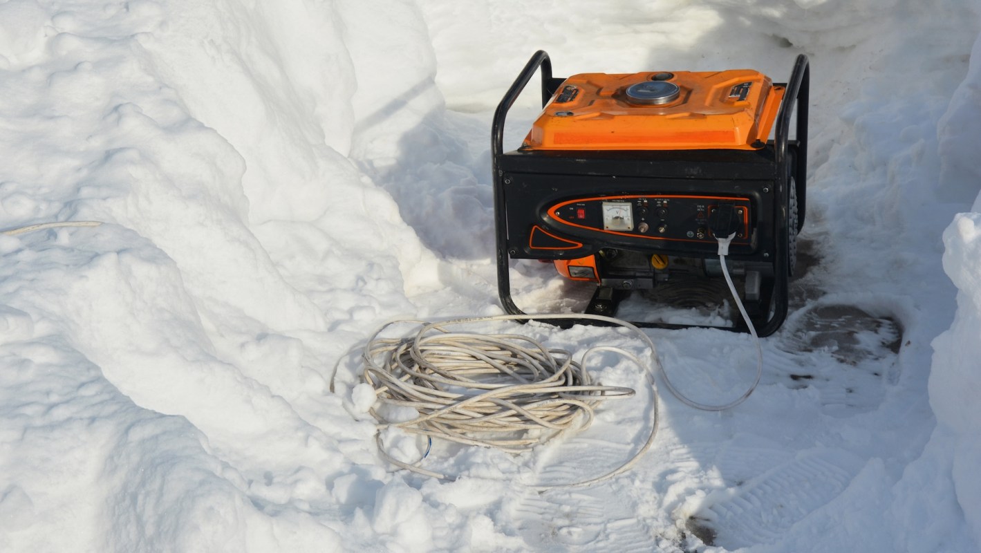 A generator sits in a snowy area next to a coiled extension cord.