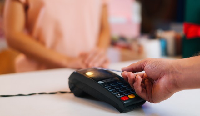 A hand holds a credit card above a payment terminal at a checkout counter.