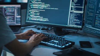 A person types on a keyboard in front of multiple computer screens displaying code.