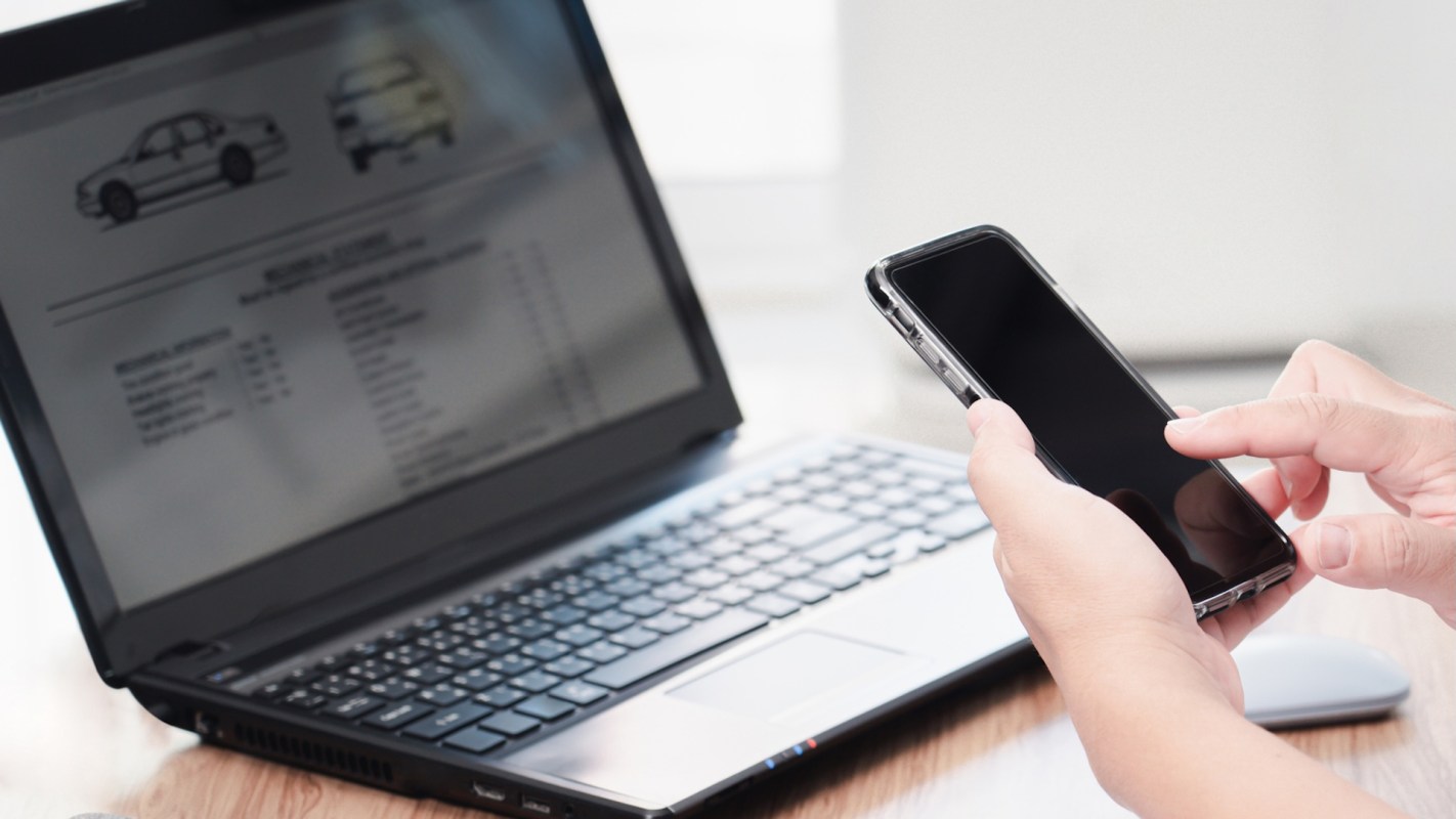 A person using a smartphone while looking at a laptop displaying car-related information.