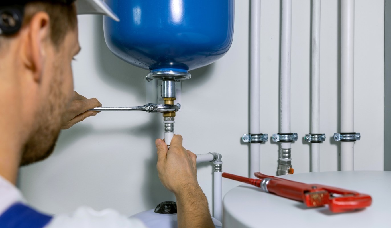 A person uses a wrench to connect piping to a blue pressure tank in a plumbing installation.