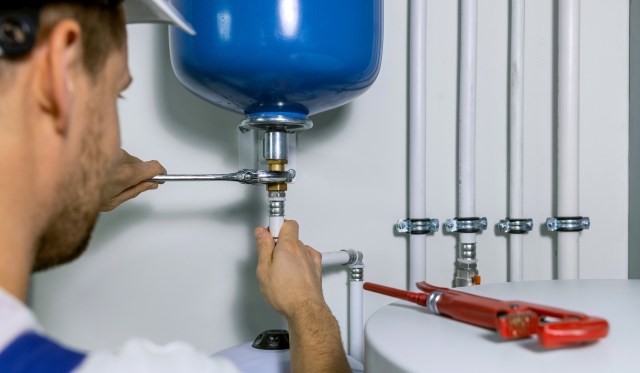 A person uses a wrench to connect piping to a blue pressure tank in a plumbing installation.