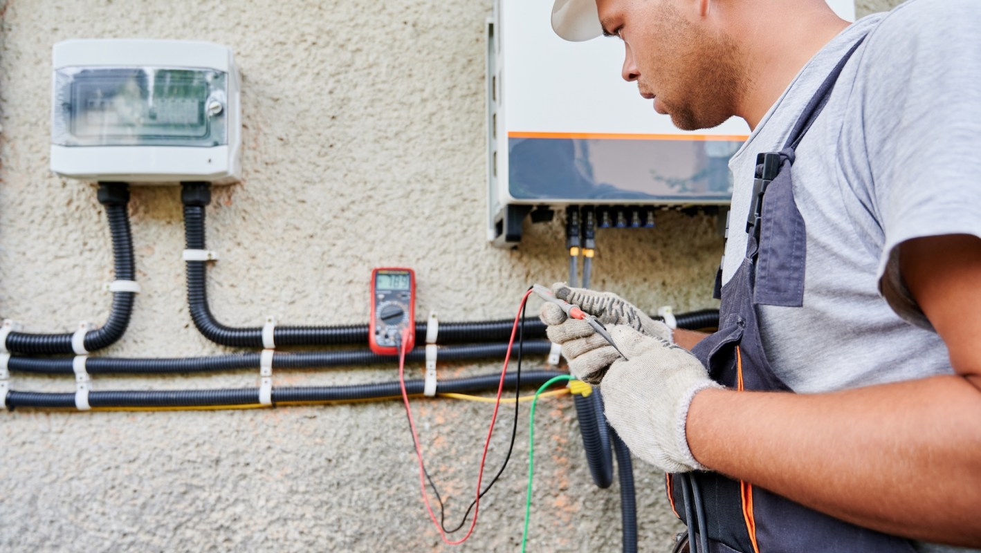 A technician uses a multimeter on electrical connections mounted on a wall.