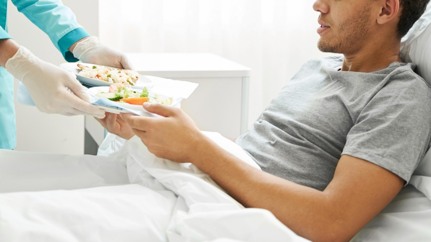 A man in a hospital bed receives a meal from a health care worker.