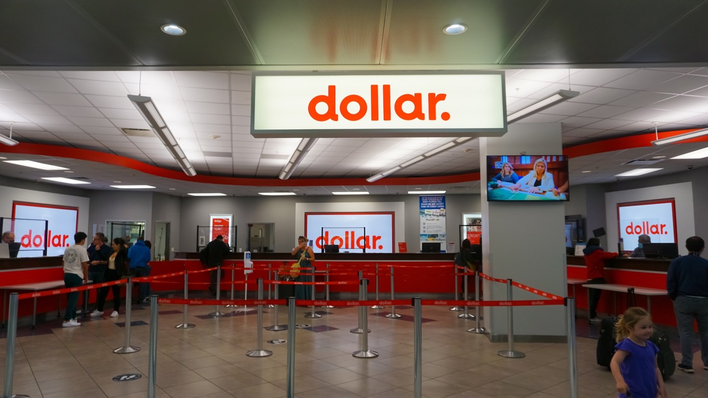 A rental car service counter with people in line and a large sign displaying the word "dollar."
