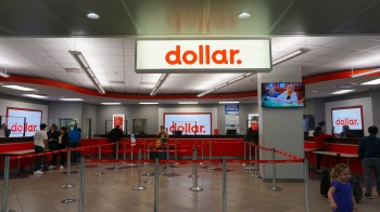 A rental car service counter with people in line and a large sign displaying the word "dollar."