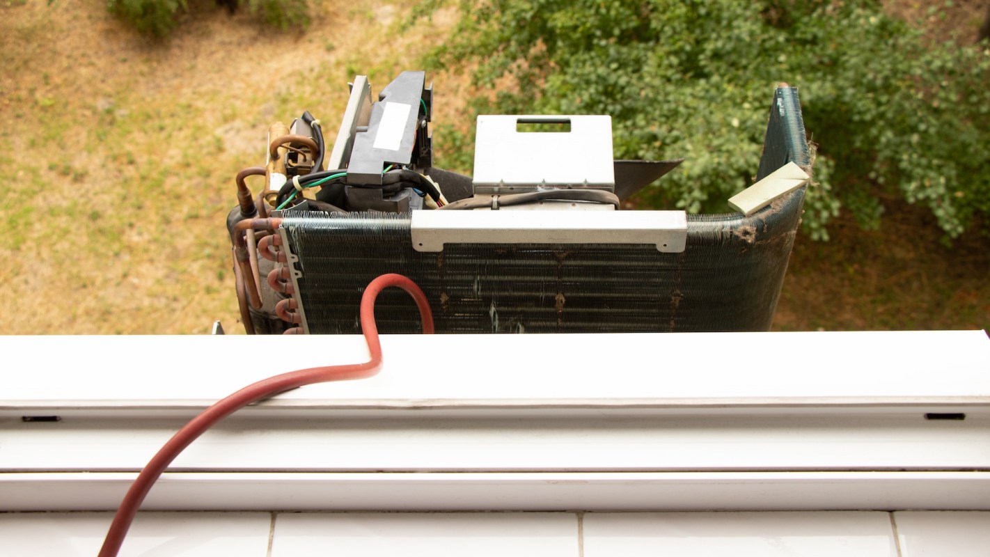 An open air conditioning unit is positioned outside a window, with greenery visible below.