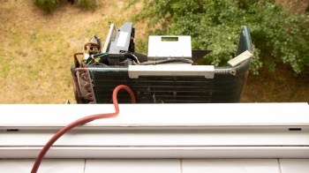 An open air conditioning unit is positioned outside a window, with greenery visible below.