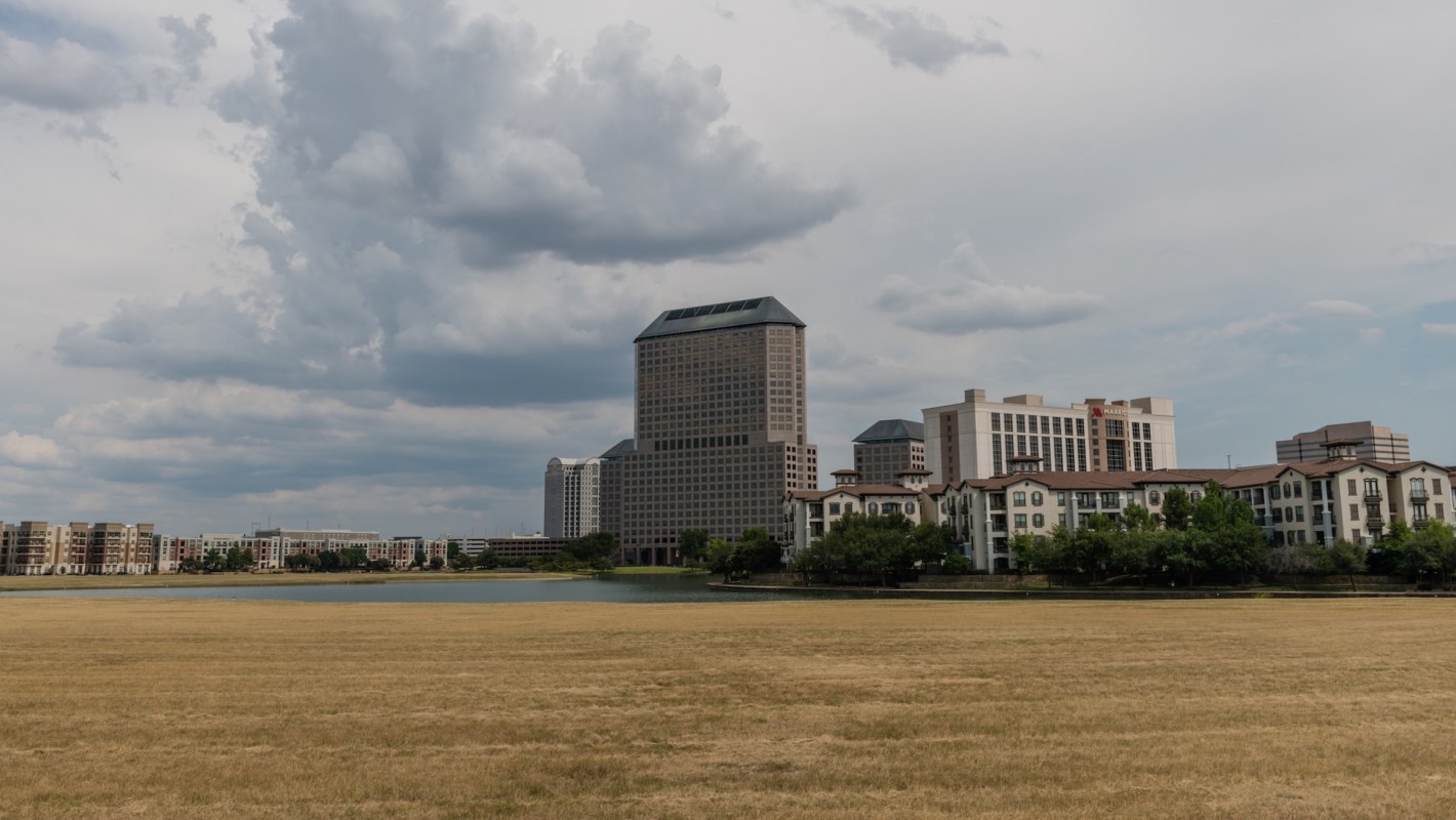A Dallas skyline featuring modern buildings near a calm lake and a field of dry grass under a cloudy sky.