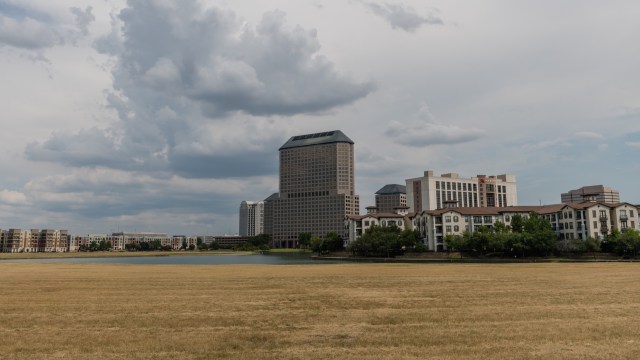 A Dallas skyline featuring modern buildings near a calm lake and a field of dry grass under a cloudy sky.