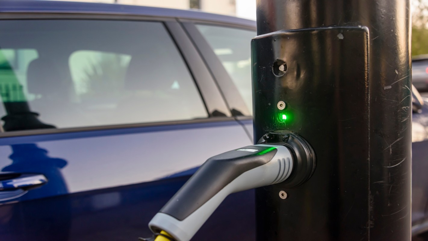 An electric vehicle charging station with a charging cable and indicator light, next to a parked car.