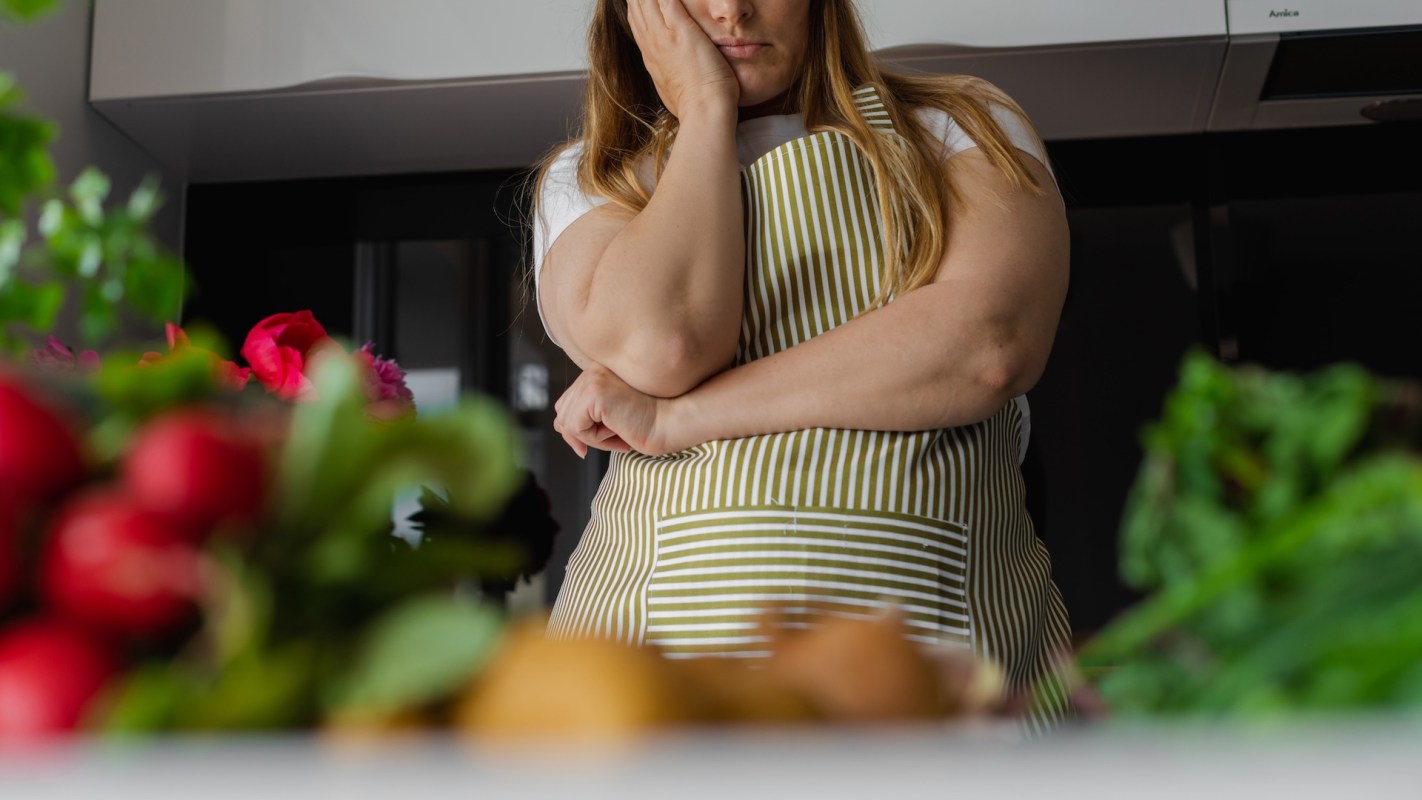 A woman stands in a kitchen, surrounded by vegetables and flowers, with her hand on her face.