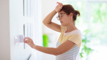 A woman adjusts a thermostat with her hand on her forehand while standing in a bright, indoor space with plants.