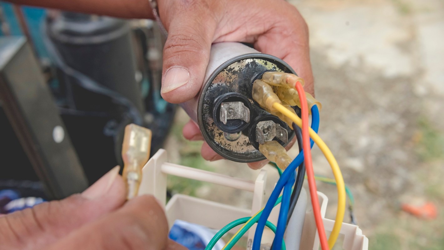 A hand holds a capacitor with multiple colored electrical wires attached.