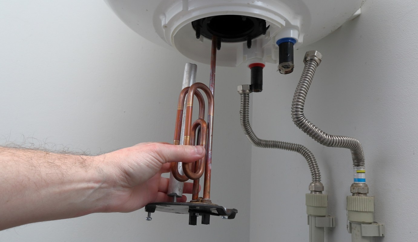 A hand holding a copper heating element under a white water heater.