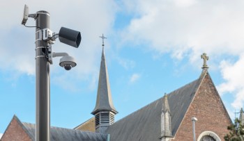 A surveillance camera and speaker mounted on a pole next to a church with a tall steeple.