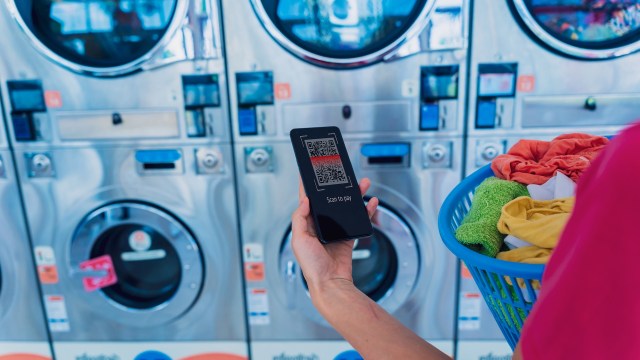 A person holds a smartphone displaying a QR code in a laundromat with washing machines in the background.