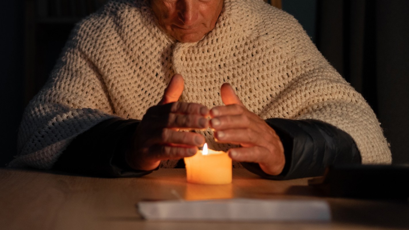 A senior citizen sitting at a table with a lit candle, hands placed above it, wearing a knitted blanket.