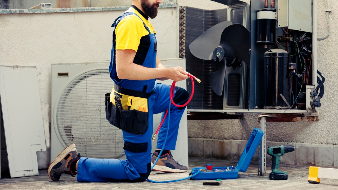 A technician kneels beside an open air conditioning unit, holding a red hose and tools.