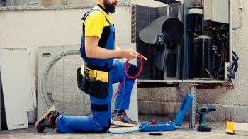A technician kneels beside an open air conditioning unit, holding a red hose and tools.