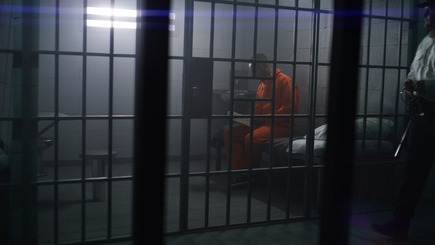 A person in an orange jumpsuit sits in a jail cell with a guard nearby.