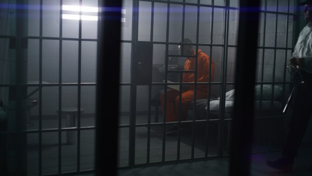 A person in an orange jumpsuit sits in a jail cell with a guard nearby.
