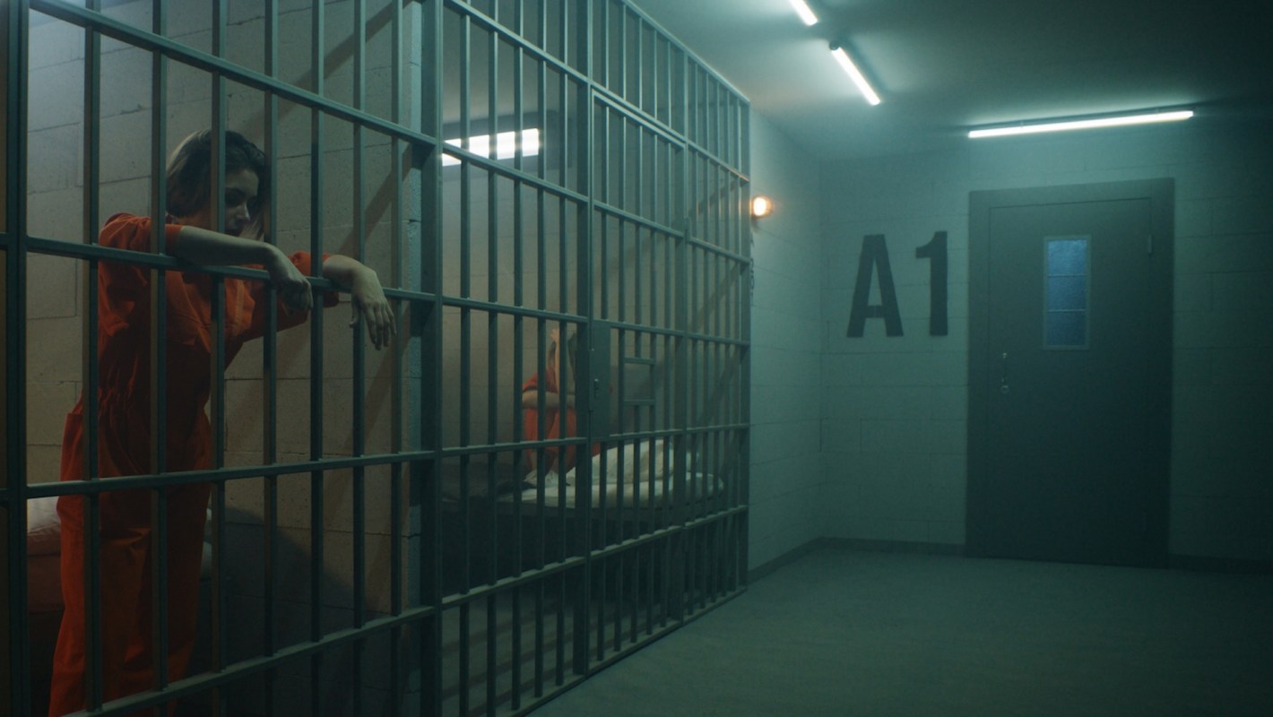 A woman in an orange jumpsuit leans on the bars of a prison cell.