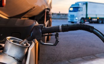A diesel fuel nozzle is positioned over a fuel tank, with a truck in the background.