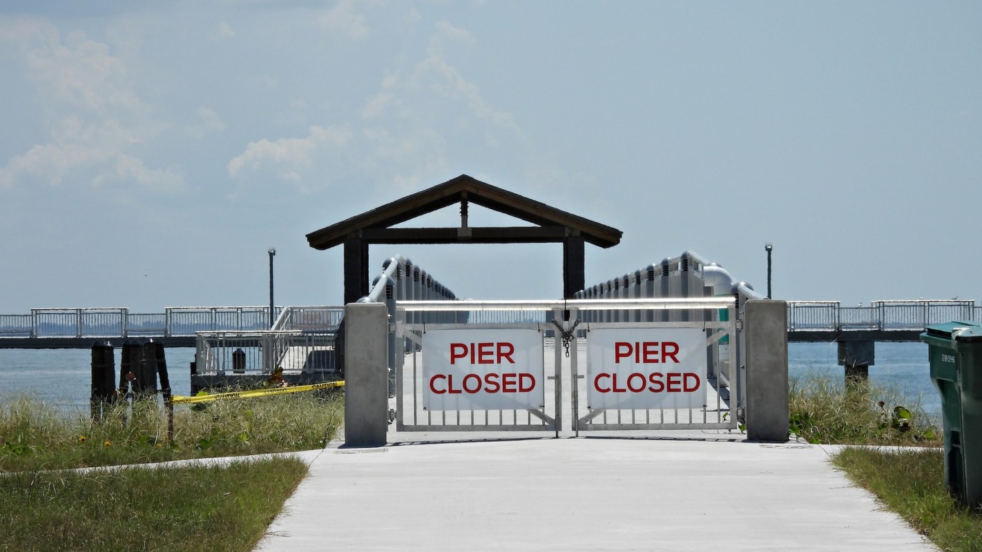 A closed pier entrance with gates displaying "PIER CLOSED" signs and a view of the water in the background.