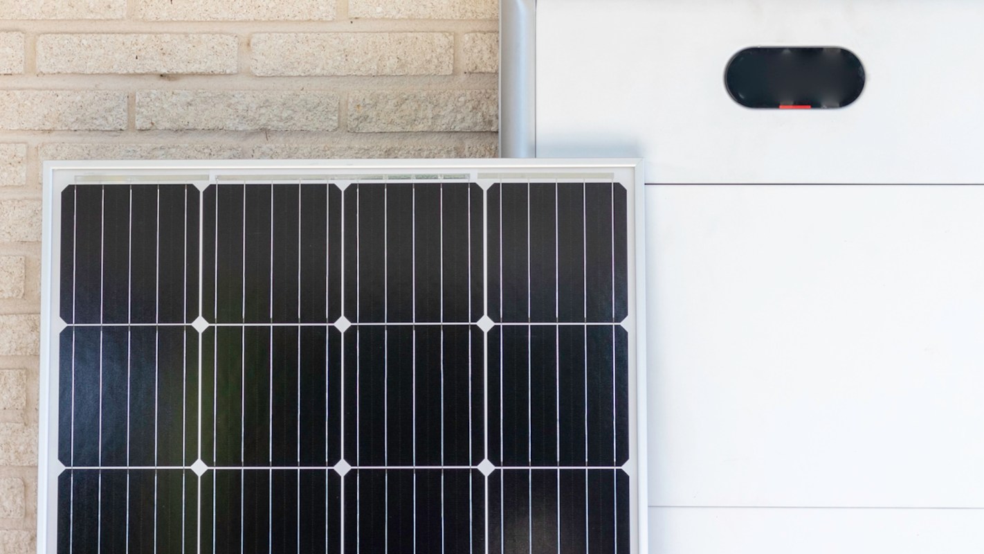 A solar panel mounted next to a white battery storage unit against a brick wall.