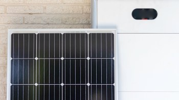 A solar panel mounted next to a white battery storage unit against a brick wall.