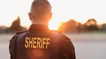 A sheriff stands with their back to the camera, facing a sunset in a rural setting.