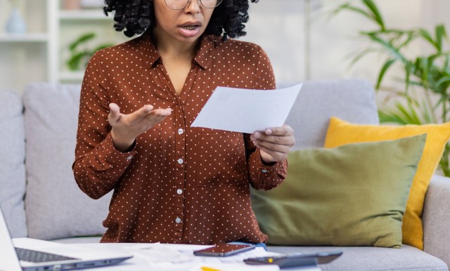 A dark-skinned woman looks at a document with a concerned expression while sitting on a couch with cushions.