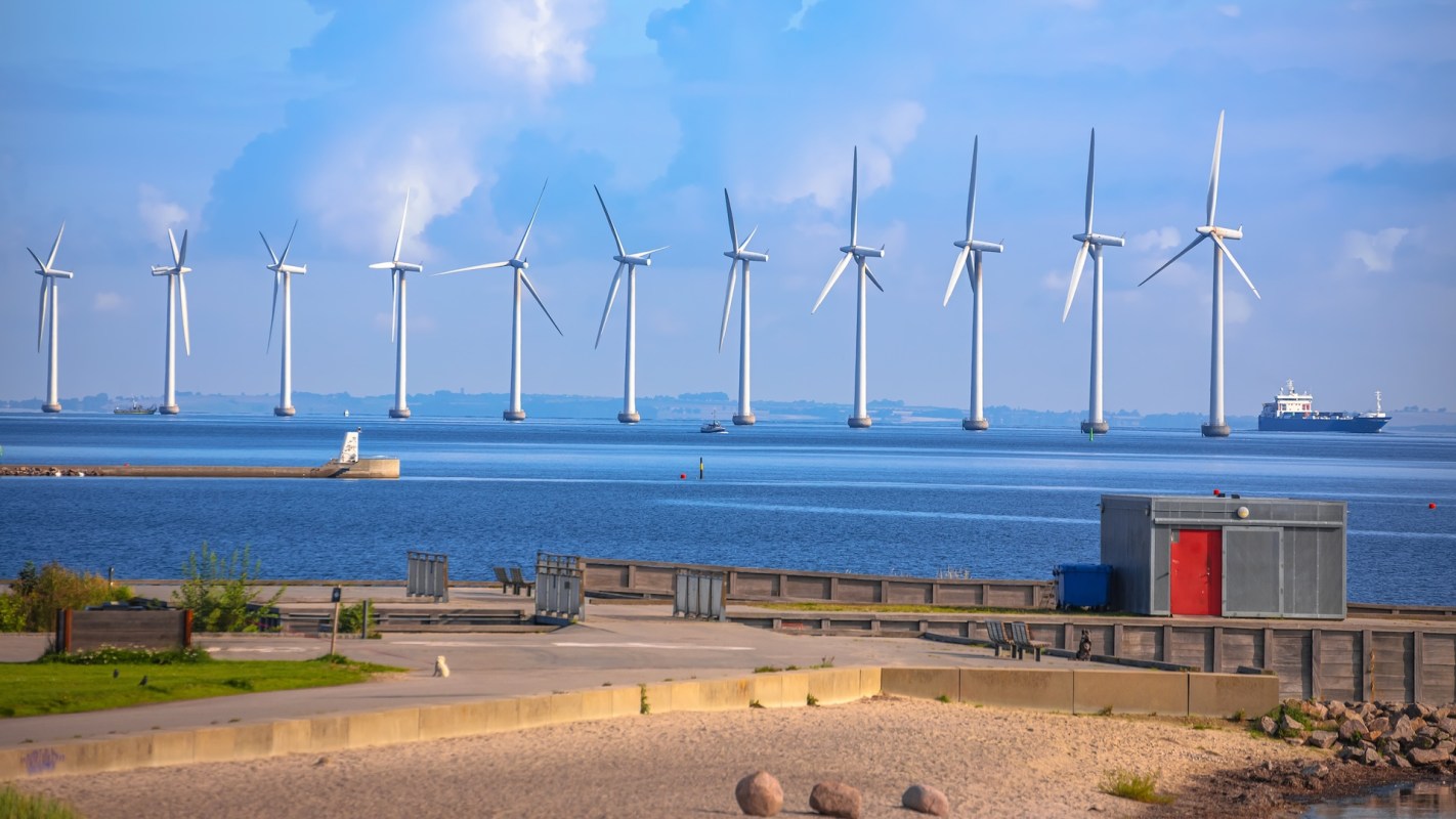 A row of wind turbines stands in a body of water with a distant boat and a shoreline structure.