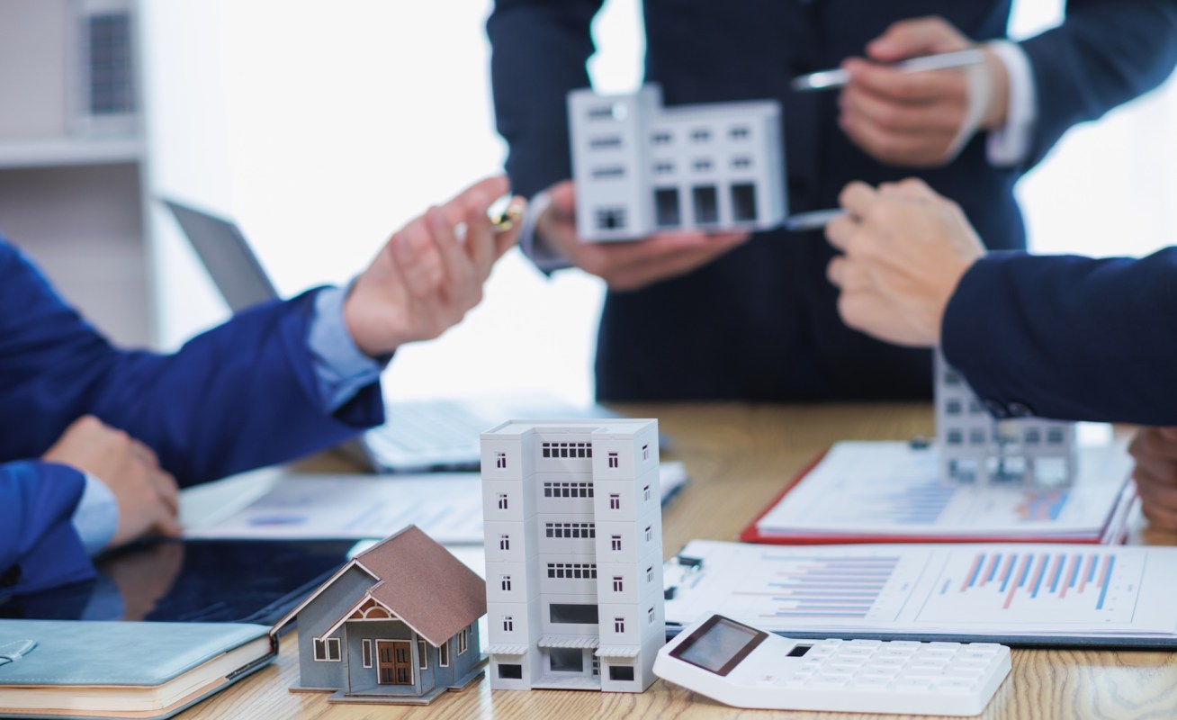Three people discuss real estate using mini models of buildings on a table with charts and a calculator.