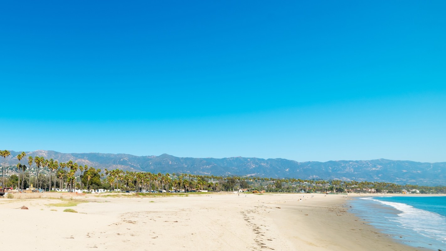 A sandy beach lined with palm trees and mountains in the background under a clear blue sky.