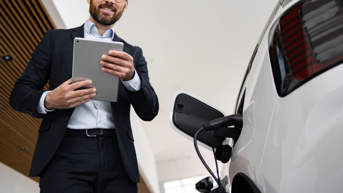 A man holding a tablet stands next to a charging electric vehicle.
