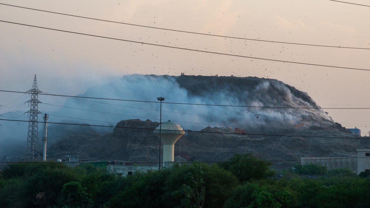 A large waste mound in a landfill emits smoke in the distance with power lines and vegetation in the foreground.