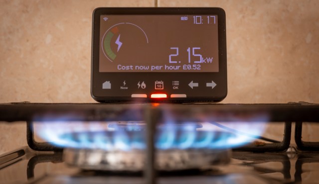 A digital energy monitor displays usage and cost above a gas burner with a blue flame.