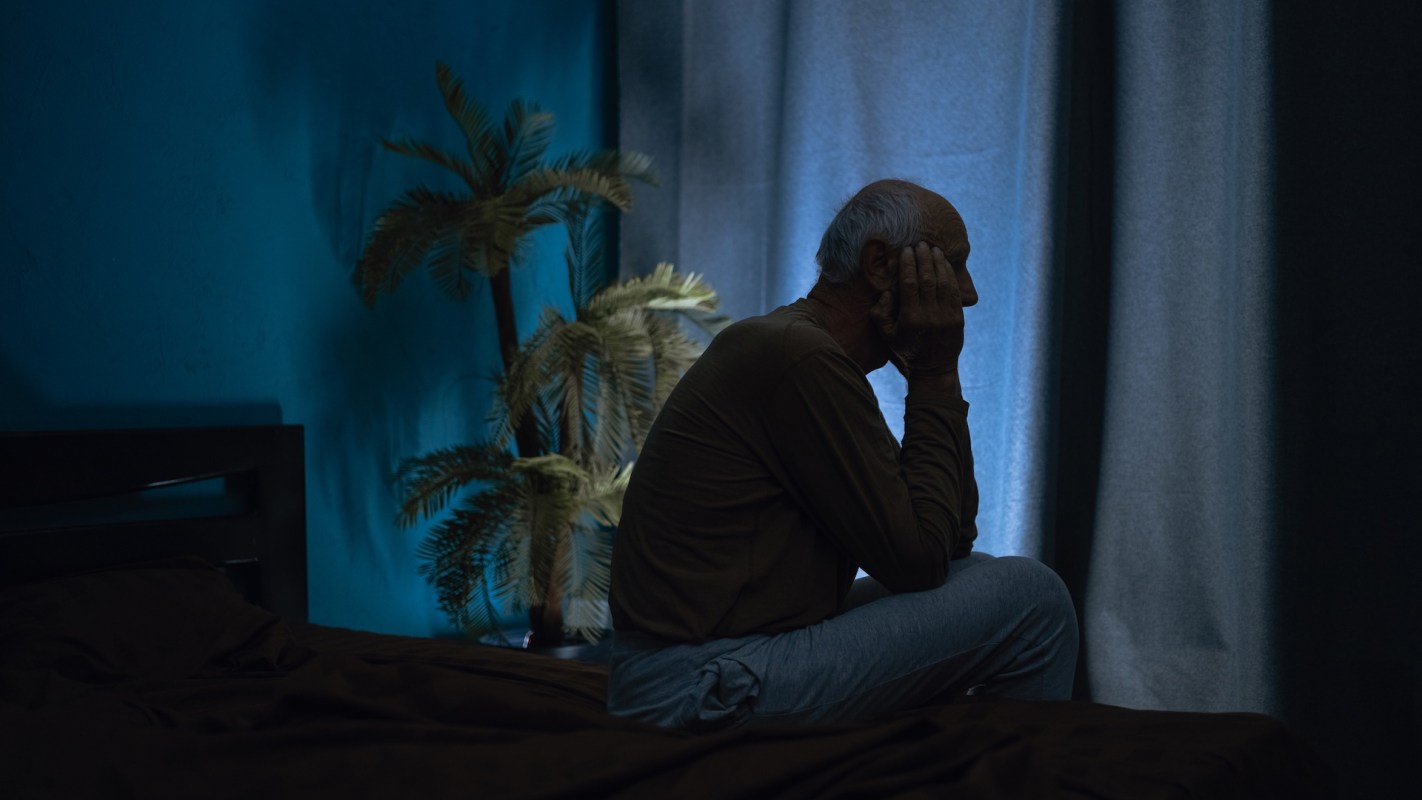 An older man sits on a bed with his head in his hands, next to a potted plant and blue curtains.