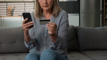 An elderly woman holds a phone in one hand and a credit card in the other while seated on a couch.
