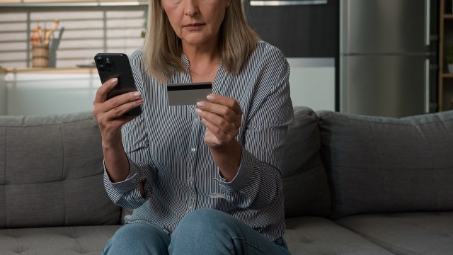 An elderly woman holds a phone in one hand and a credit card in the other while seated on a couch.